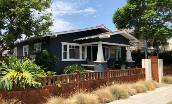 dark blue bungalow style home on a sunny street behind a low, wooden fence