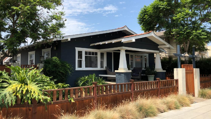 dark blue bungalow style home on a sunny street behind a low, wooden fence